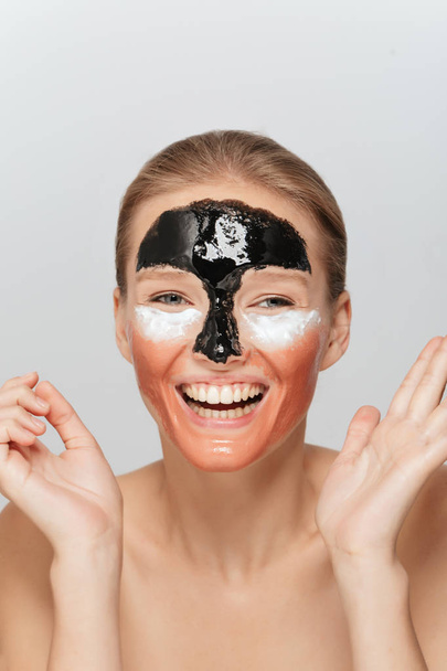 Portrait of young cheerful woman with different cosmetic masks on face joyfully looking aside over gray background - Photo, Image