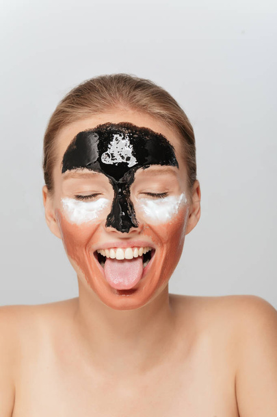 Ritratto di giovane donna gioiosa con diverse maschere cosmetiche sul viso felicemente chiudendo gli occhi e mostrando la lingua sulla macchina fotografica su sfondo grigio
 - Foto, immagini