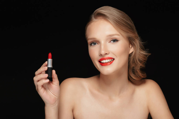 Portrait of young beautiful woman with wavy hair holding red lipstick in hand happily looking in camera over black background - Photo, Image