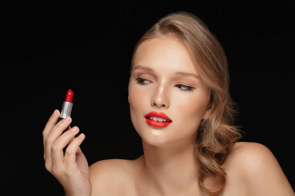 Portrait de jeune femme séduisante aux cheveux ondulés regardant avec rêverie le rouge à lèvres rouge sur fond noir
 - Photo, image