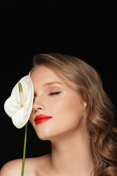 Portrait of young attractive lady with wavy hair and red lips dreamily covering eye with white calla flower over black background - Photo, Image