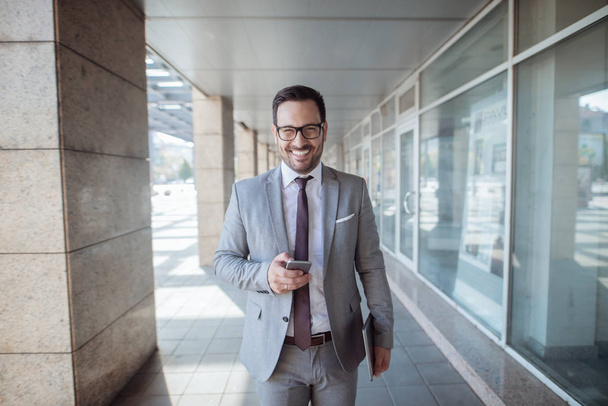 Улыбающийся бизнесмен, держащий смартфон в одной руке и планшет в другой, стоя перед бизнес-центром
. - Фото, изображение
