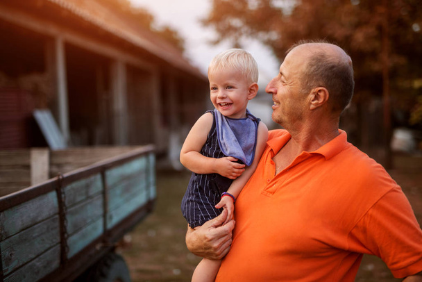 Bild eines stolzen Großvaters, der seinen Enkel hält und ihn mit Liebe betrachtet. im Hinterhof eines Bauernhofes stehen. - Foto, Bild