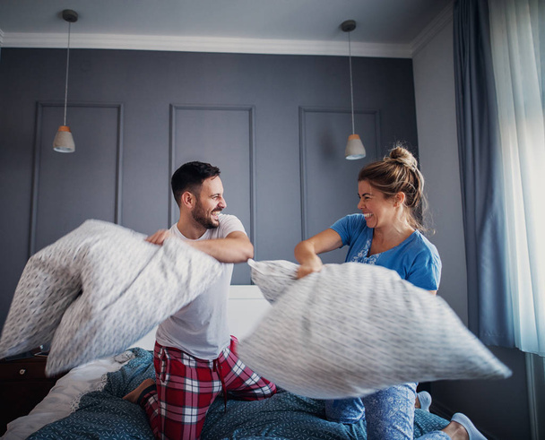 Beautiful young woman and man pajamas having fun in bedroom and fighting with pillows  - Photo, Image