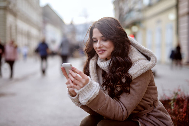 Menina bonito em casaco de inverno de pé na rua e olhando para o telefone. Sorrindo e parecendo feliz
. - Foto, Imagem