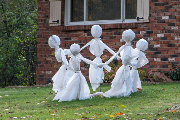 Un círculo de fantasmas vestidos de blanco juegan un juego de anillo alrededor de la rosie mientras bailan en el césped de una casa decorada para Halloween
 - Foto, imagen