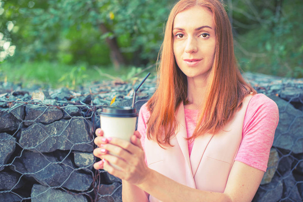 A beautiful red-haired girl goes down the stairs and drinks coffee from a paper Cup. Lifestyle Breakfast in the Park - Foto, Bild