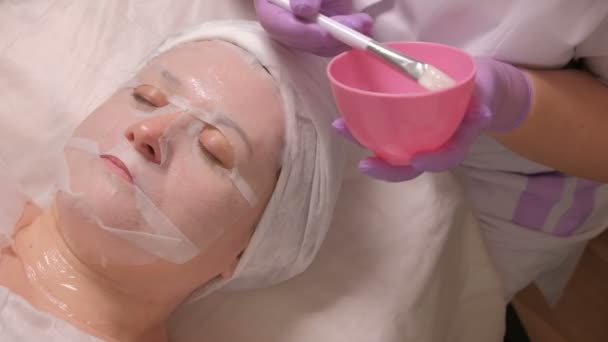 Kobieta na procedury odmładzania skóry w centrum kosmetologii. Kosmetyczka z pomocą biały pędzel stosuje żel przez maskę na twarz kobiety w gabinecie kosmetycznym. Sesja terapii medycznej. - Materiał filmowy, wideo
