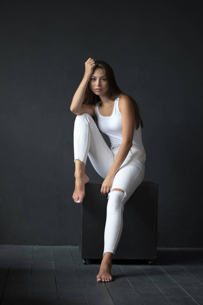 Beautiful woman in white tank-top and jeans is sitting on a black background. - Фото, зображення