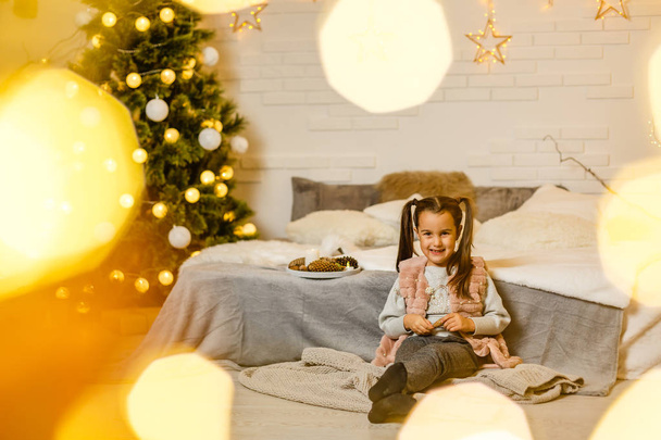 agradable niña encantadora apoyada en la cama mientras está sentada en el suelo y mirando a la cámara cerca del árbol de Navidad en el interior brillante de la casa
 - Foto, imagen