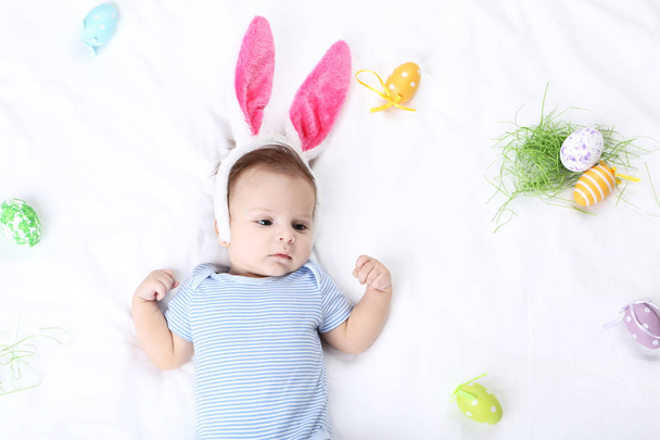 Cute baby with easter eggs and rabbit ears lying on white bed - Foto, Imagem