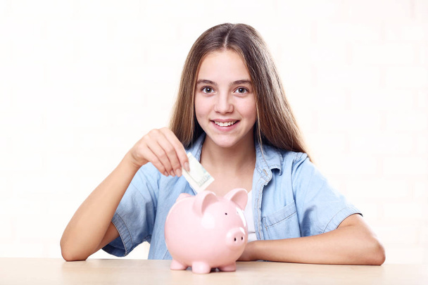 Young girl with pink piggybank and dollar banknote sitting at table - Foto, afbeelding