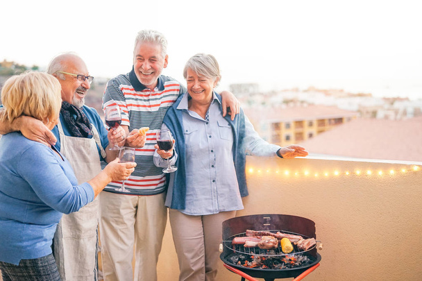 Heureux amis aînés qui s'amusent à boire du vin rouge au barbecue dîner en terrasse - Les personnes âgées dînent et rient ensemble sur le toit - Concept d'amitié et de mode de vie des personnes âgées
 - Photo, image