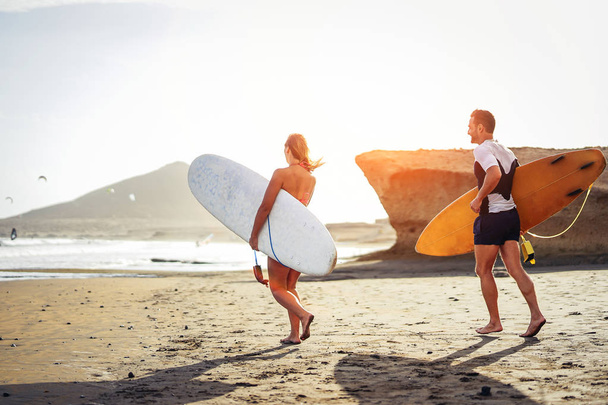 Pareja de surfistas corriendo junto con tablas de surf en la playa al atardecer - Amigos deportivos divirtiéndose yendo a surfear - Viajes, vacaciones, deporte concepto de estilo de vida
 - Foto, Imagen