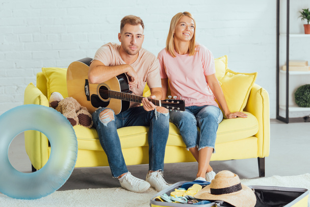 husband holding guitar while wife dreaming about summer holidays and sitting on couch at home - Photo, Image