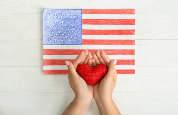 Mulher segurando coração vermelho perto do desenho da bandeira nacional americana na mesa de madeira branca
 - Foto, Imagem