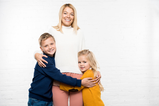 glückliche Mutter umarmt Sohn und Tochter und schaut in die Kamera auf weißem Hintergrund - Foto, Bild