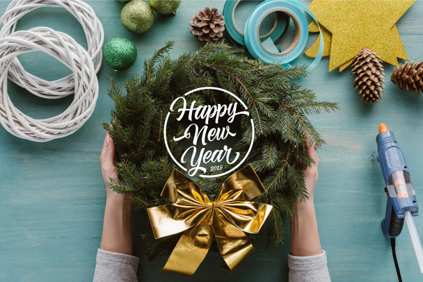 tiro recortado de mujer sosteniendo coronas de pino hechas a mano con arco de oro y decoraciones en la mesa de madera azul con letras de "feliz año nuevo"
 - Foto, Imagen