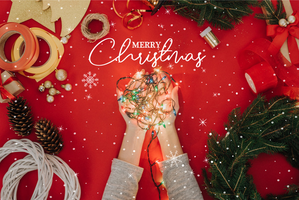 cropped shot of woman holding christmas lights in hands with pine tree branches and decorations for handmade wreath on red backdrop with "merry christmas" lettering - Photo, Image