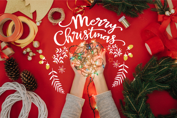 松の木の枝と「メリー クリスマス」の文字と赤い背景の手作りリース飾り手にクリスマス ライトを保持している女性のクロップ撮影 - 写真・画像