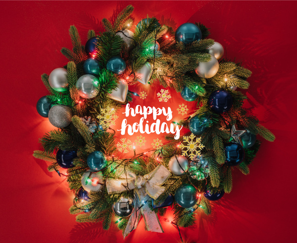 bovenaanzicht van de kroon van Kerstmis versierd met speelgoed en lichten op rode achtergrond met "happy holiday" belettering - Foto, afbeelding
