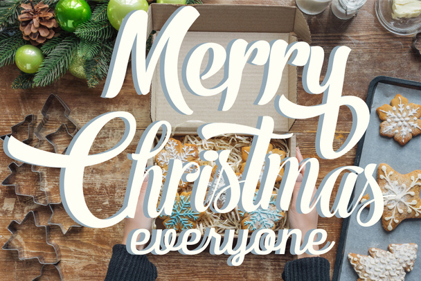 cropped shot of woman holding box with  homemade christmas cookies on wooden tabletop with decorative christmas wreath with "merry christmas everyone" lettering - Фото, зображення