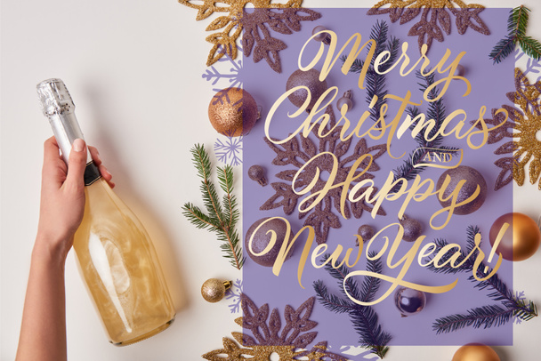 cropped image of woman holding bottle of sparkling champagne near christmas decoration on white with "merry christmas and happy new year" lettering - Photo, Image