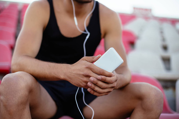 Sportsman listening music sitting on stadium seats, relax after workout. Focus on smartphone - Fotó, kép
