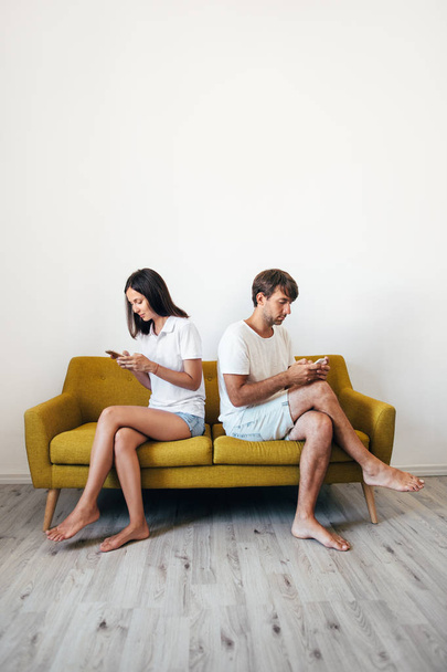 Young couple sitting on sofa back to back using mobile phones. Problem in relationship - 写真・画像