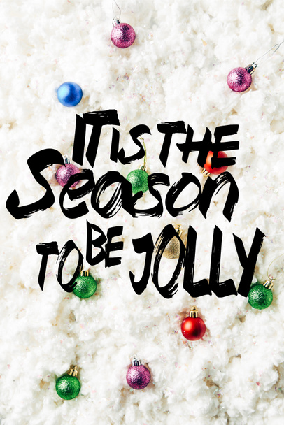 top view of colorful christmas toys on white cotton wool background with "it is the season to be jolly" inspiration - Fotó, kép