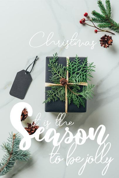 top view of black christmas gift box with fir branches and tag on marble table with "christmas - it is the season to be jolly" inspiration  - Фото, изображение