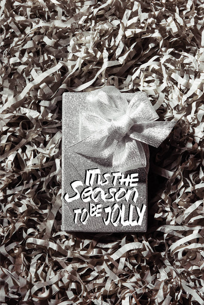 top view of silver christmas gift on grey paper confetti backdrop with "it is the season to be jolly" inspiration  - Фото, изображение