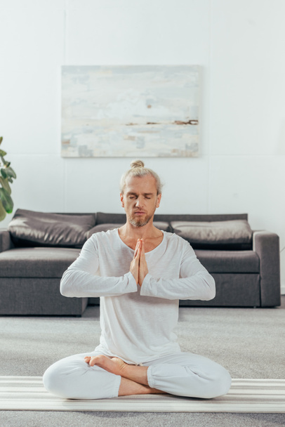 adult man with closed eyes meditating in lotus position and namaste gesture at home  - Foto, Bild