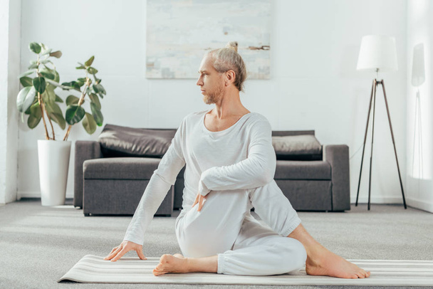 skoncentrowane dorosły człowiek siedzi w jogi maty i odwracając w domu - Zdjęcie, obraz
