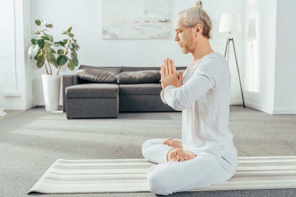 Seitenansicht des meditierenden Mannes in Lotusposition mit namaste Geste zu Hause   - Foto, Bild