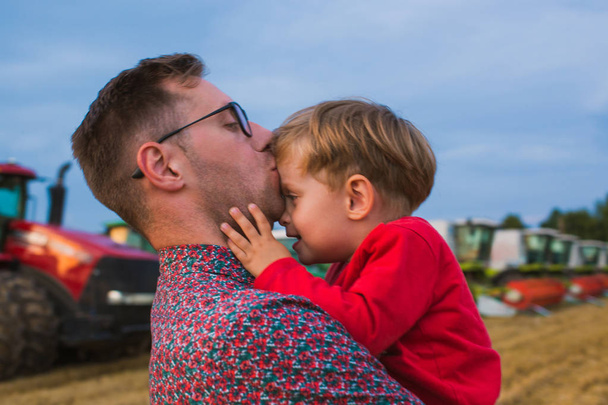 Close-up o pai feliz segurando seu filhinho nos braços e beijando-o contra o fundo dos tratores e combina no campo
 - Foto, Imagem