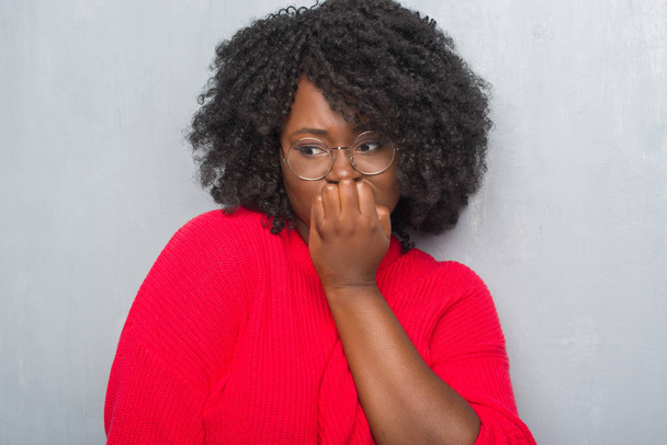 junge afrikanisch-amerikanische Frau über der grauen Grunge-Wand mit Winterpullover und Brille, die gestresst und nervös aussieht, die Hände auf mundgebissenen Nägeln. Angstproblem. - Foto, Bild