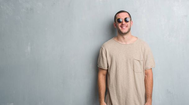 Молодой кавказский мужчина на серой гранж-стене в солнечных очках со счастливой и прохладной улыбкой на лице. Счастливчик
. - Фото, изображение