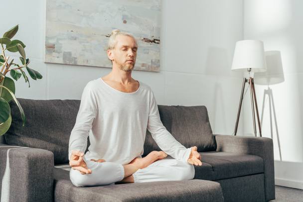 Erwachsener Mann mit geschlossenen Augen meditiert in Lotusposition auf dem heimischen Sofa - Foto, Bild
