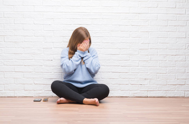 Giovane donna adulta seduta sul pavimento in autunno su un muro di mattoni bianchi con espressione triste che copre il viso con le mani mentre piange. Concetto di depressione
. - Foto, immagini