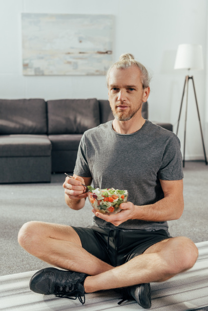 hombre atlético en ropa deportiva sosteniendo un tazón de ensalada de verduras y mirando a la cámara mientras está sentado en la esterilla de yoga en casa
  - Foto, Imagen