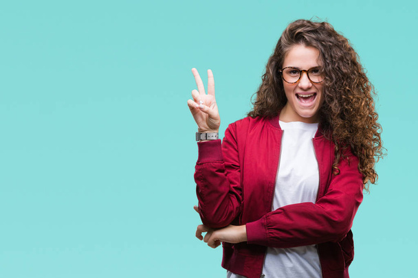 Beautiful brunette curly hair young girl wearing jacket and glasses over isolated background smiling with happy face winking at the camera doing victory sign. Number two. - Photo, Image