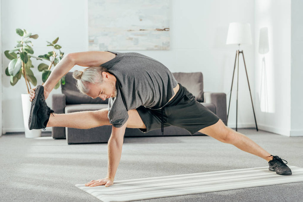 athletic adult man in sportswear stretching on yoga mat at home  - Fotografie, Obrázek