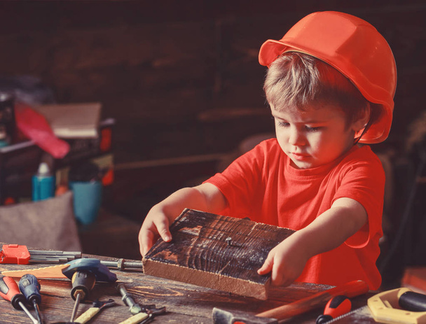 Child in helmet cute playing as builder or repairer, repairing or handcrafting. Handcrafting concept. Kid boy hammered nail into wooden board. Toddler on busy face plays at home in workshop - 写真・画像