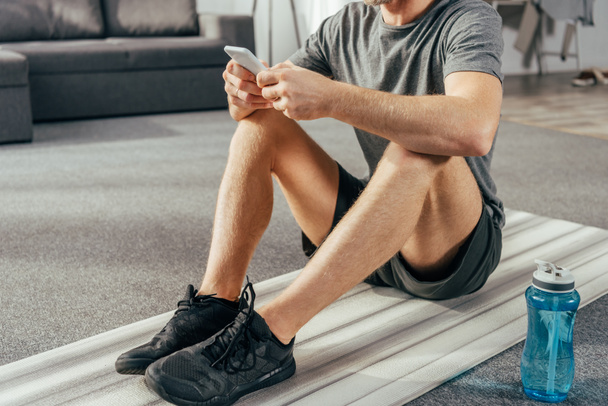 cropped shot of athletic man in sportswear sitting on yoga mat and using smartphone at home - Foto, Imagen