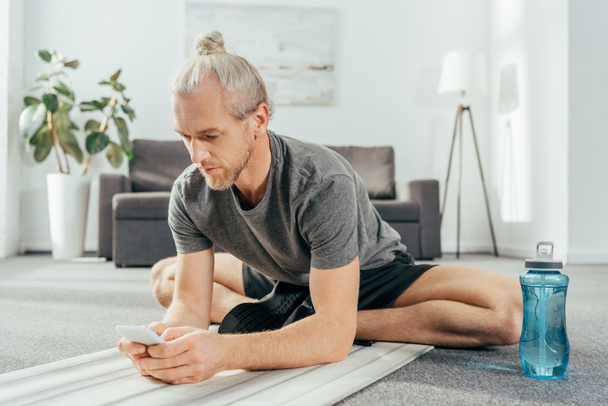 hombre adulto deportivo sentado en la esterilla de yoga y el uso de teléfono inteligente en casa
 - Foto, Imagen