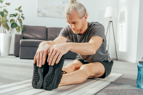 athletischer erwachsener Mann in Sportbekleidung zu Hause auf Yogamatte sitzend und streckend - Foto, Bild