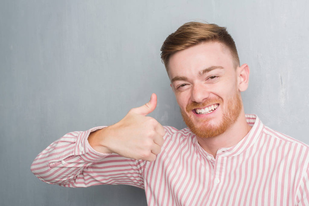 Jonge roodharige man over grijs grunge muur blij met de grote glimlach doen ok teken, duim omhoog met vingers, uitstekende teken - Foto, afbeelding