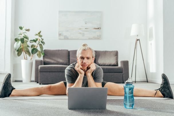 sporty adult man doing split on yoga mat and looking at laptop - Zdjęcie, obraz