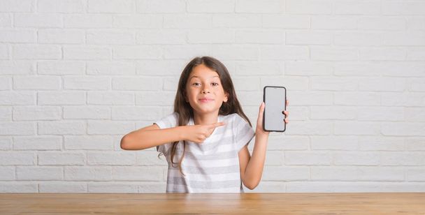 Giovane bambino ispanico seduto sul tavolo utilizzando smartphone molto felice puntando con mano e dito
 - Foto, immagini
